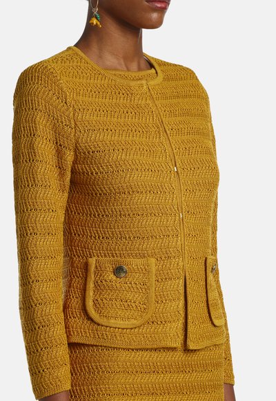 Frau trägt eine senfgelbe, strukturierte Strickjacke mit rundem Ausschnitt, langen Ärmeln und vorne aufgesetzten Taschen mit Knöpfen, kombiniert mit einem passenden Rock.