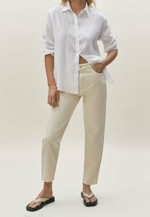 Frau trägt ein lockeres weißes Hemd mit Knöpfen, beige gerade geschnittene Hosen und beige Flip-Flops mit schwarzen Sohlen, steht mit einer Hand in der Tasche.