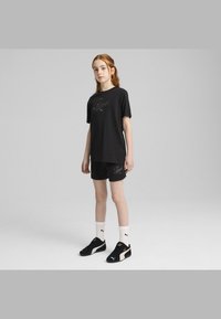 Camiseta de algodón negra con el logo de "Puma", combinada con pantalones cortos negros que presentan el mismo logo. Lleva puesto unas zapatillas negras y doradas y calcetines blancos.