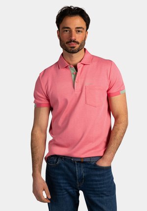 Man die een roze poloshirt met korte mouwen en een borstzak draagt, samen met een donkerblauwe spijkerbroek, staand met één hand in de zak.