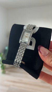 Sidabrinis laikrodis su stačiakampiu ciferblatu, baltu skaitmeniniu paviršiumi, auksiniais valandų ženklais, deimantų pavidalo akcentais ant rėmelio ir metaline grandinėlės apyranke, pateiktas ant juodos pagalvėlės.