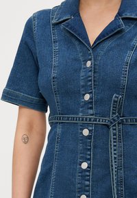 Δερμάτινο φόρεμα με γιακά πουκαμίσου, κοντά μανίκια, κλείσιμο με κουμπιά εμπρός, ζώνη στη μέση και σκούρο μπλε χρώμα με αντίθετες ραφές.