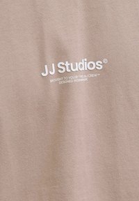 Tricou bej cu o textură moale din bumbac, cu un text alb imprimat: "JJ Studios", "Făcut pentru tine de echipa J&J" și "Proiectat în Danemarca."