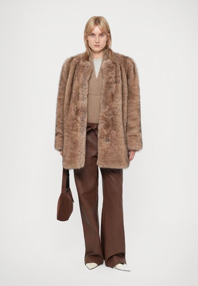 Abrigo de piel sintética marrón con un corte holgado, que presenta mangas largas y un collar. Combinado con una blusa de punto beige y pantalones de pierna ancha marrón oscuro.