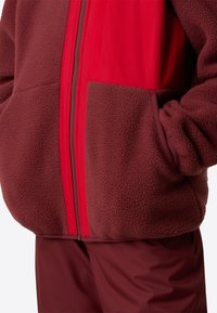 Κόκκινο και μαρόν fleece μπουφάν με φερμουάρ στο μπροστινό μέρος, διαθέτοντας υφή στην επιφάνεια και πλαϊνές τσέπες. Το σχέδιο περιλαμβάνει ομαλή εσωτερική επένδυση.