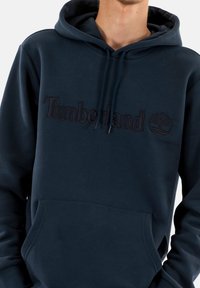 Marineblauwe hoodie van katoen, met een geborduurd "Timberland" logo, een capuchon met trekkoorden en een voorzak in kangoeroestijl.