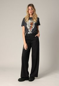 Pantalons larges noirs à taille haute, associés à un t-shirt gris foncé arborant un graphique de tigre et une écriture audacieuse. Des chaussures noires complètent le look.