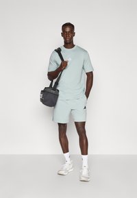 Gaiši zils sporta t-krekls un šorti no gludas auduma. Modelis tur melnu Adidas somu ar baltu logotipu. Balti sporta apavi un zeķes.