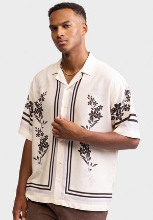 Uomo che indossa una camicia crema a maniche corte con motivi floreali neri e righe, in piedi con un'espressione neutra contro uno sfondo semplice.