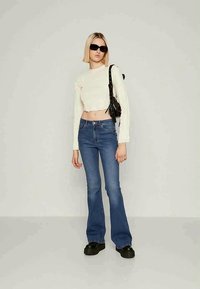Top cropped branco de manga longa, jeans azuis de perna larga e sapatos de plataforma pretos. Óculos de sol e uma pequena bolsa preta completam o look.