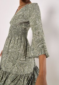 Long-sleeve dress with a green paisley pattern, smocked waist, V-neckline, and gathered hem. Made of lightweight fabric with a soft texture.