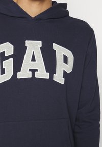 Person som bär en marinblå huvtröja med stora vita bokstäver "GAP" över bröstet, med en framficka och huva.