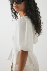 Witte blouse met pofmouwen en peplumontwerp, gemaakt van een gladde stof. Model draagt ronde, schildpadbril. Lichte achtergrond.