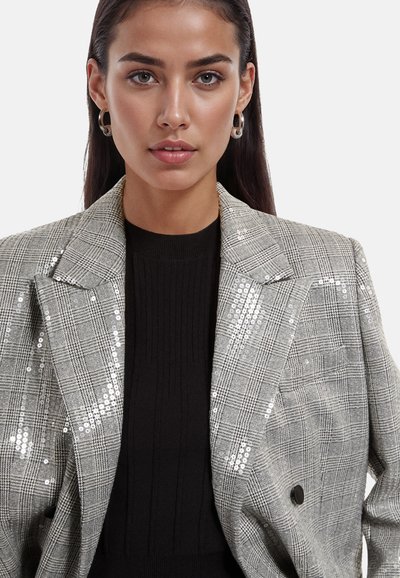 Blazer à carreaux gris avec paillettes, design à double boutonnage, rabats aigus et pull noir texturé en dessous. Boucles d'oreilles en argent en forme de créole.
