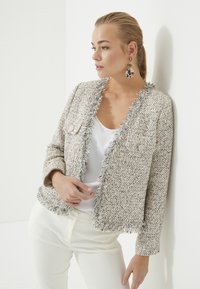 Geverfde, lichtgekleurde tweedjas met franjes aan de randen, voorzien van twee voorzakken en knoopsluitingen, gedragen over een witte top en lichte broek.