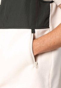 Veste en polaire blanche avec une section supérieure noire, dotée d'une poche zippée sur le côté et d'une texture lisse. Le tire-zipp est métallique.