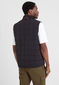 Black quilted vest with high collar and orange accent on the back, worn over a white shirt. Textured material and sleeveless design.