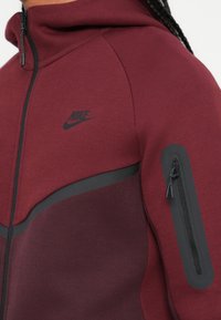 Casaco desportivo em bordeaux com um logo da Nike em preto, com fecho de correr, capuz e bolso lateral com fecho. Feito de um tecido liso e texturizado.