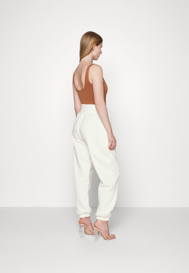 Bodysuit marrón sin mangas, pantalones de chándal blancos de corte relajado con puños elásticos, sandalias transparentes de tacón. Diseño sencillo, textura de tela suave.