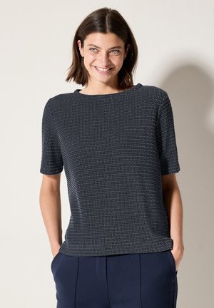 Marineblauwe top met korte mouwen met een textuurpatroon, ronde halslijn en een relaxte pasvorm. Draag met marineblauwe broek, met de handen in de zakken.