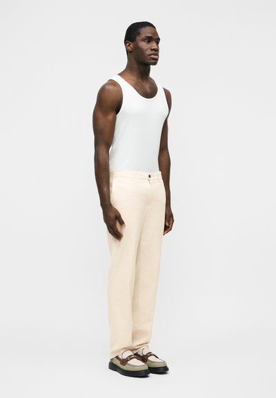 Άνδρας που στέκεται φορώντας λευκή αμάνικη μπλούζα, μπεζ παντελόνι και πράσινα και καφέ μοκασίνια, μπροστά σε απλό φωτεινό φόντο.