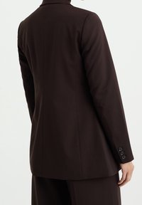Hnědý upravený blazer s hladkou texturou, strukturovaným designem, jedním rozparkem a knoflíkovými manžetami, zobrazený ze zadní strany.