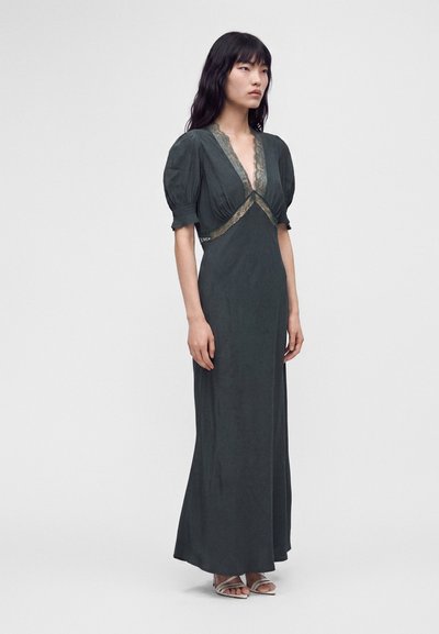 Femme portant une longue robe vert foncé avec manches bouffantes et bordure en dentelle, debout devant un fond clair uni, tournée vers la gauche.