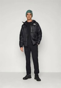 Άντρας με μαύρο μπουφάν North Face, μαύρο παντελόνι, μαύρα παπούτσια και πράσινο πλεκτό καπέλο, που στέκεται μπροστά από απλό ανοιχτό φόντο.