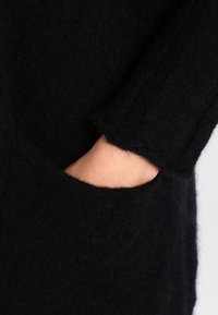Černý pletený svetr s měkkou, načechranou texturou, s rukou sahající do boční kapsy a ukazující uvolněný, oversized design.