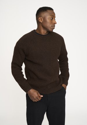 Brune ribbet sweater med lange ærmer og rund halsudskæring, lavet af en struktureret strikstof. Parret med sorte bukser, hænderne hviler langs siden.