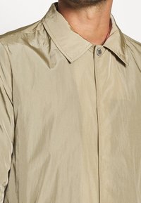 Beige lättviktsjacka med en synlig lyster, med klassisk krage och central knäppning. Strukturerad tyg med en subtil glans.