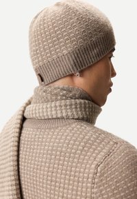 Person som bär en beige, texturerad stickad mössa, matchande halsduk och tröja, visad bakifrån och i profil med en synlig silverring i örat.