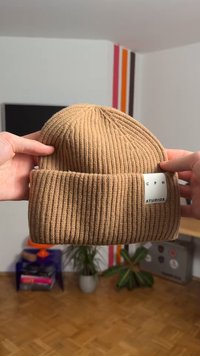 Beige ribbed knit beanie with a folded cuff, featuring a woven label with "CPH STUDIOS" text in black. Held by two hands.