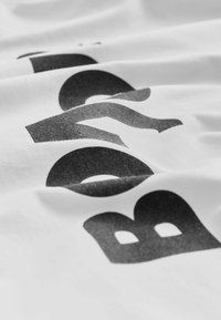 Hvid bomuldsstof med sort grafisk tekst i fed skrifttype, der viser en tekstureret finish og varierende gråtoner.