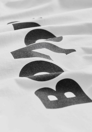 Biały bawełniany materiał z czarnym graficznym tekstem w pogrubionej czcionce, prezentujący teksturowane wykończenie i różne odcienie szarości.