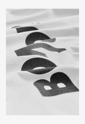 Bílá bavlněná látka s černým grafickým textem v tučném písmu, představující texturovaný povrch a různé odstíny šedé.