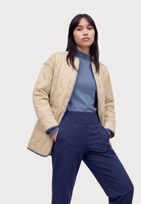 Mujer de pie con las manos en los bolsillos, lleva una chaqueta acolchada beige sobre un jersey de cuello alto azul y pantalones azul marino sobre un fondo liso.