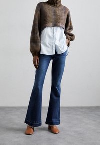 Maglione all'uncinetto marrone con colletto alto, indossato sopra una camicia bottonata azzurro chiaro; abbinato a jeans scuri a zampa d'elefante e tacchi marroni.
