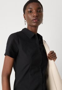 Svart kortermet skjorte med teksturert stoff, spiss krage, synlige knapper og en fitted design, holdt sammen med en lysfarget veske.