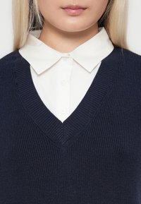 Pull en maille bleu marine avec un col en V, porté par-dessus une chemise blanche à col boutonné. Tissu texturé, coupe ajustée.