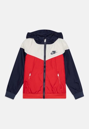 Nike windbreaker jas met capuchon, marineblauwe mouwen, wit schouderpaneel, rood lijf, rits aan de voorkant, twee met rits afsluitbare zakken en Nike-logo op de borst.