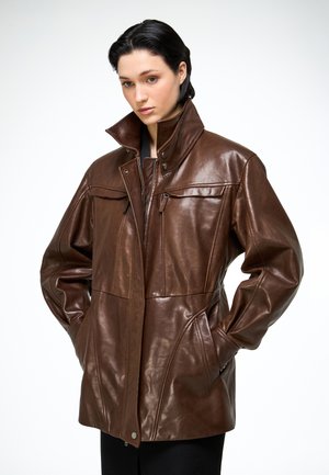 Ung person med kort mørkt hår iført en oversize brun læderjakke med hænderne i lommerne, stående mod en ensfarvet hvid baggrund.