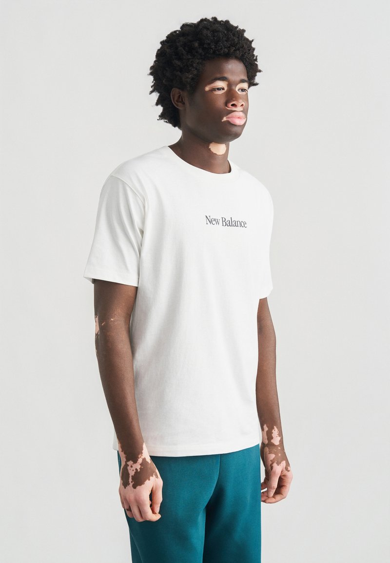 Άσπρο βαμβακερό T-shirt με το κείμενο «New Balance» σε μαύρο, λαιμόκοψη κοντό λαιμό, κοντά μανίκια, σε συνδυασμό με πετρόλ παντελόνι, φορεμένο από ανδρικό μοντέλο.