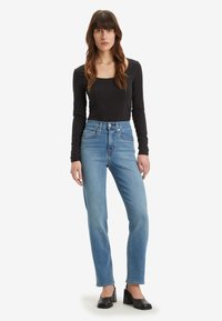 Top preto de mangas compridas com decote quadrado, combinado com jeans de cintura alta em azul claro, apresentando adornos subtis e corte reto.