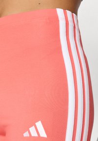 Κοραλί αθλητικά κολάν με τρεις λευκές ρίγες στο πλάι και λευκό λογότυπο κοντά στο στρίφωμα. Φτιαγμένα από λείο, ελαστικό υλικό.