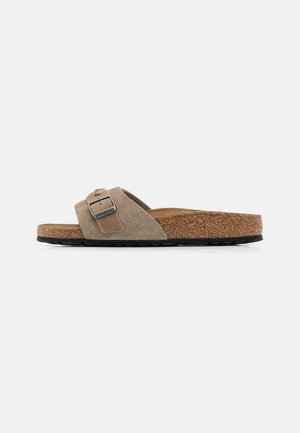 Suede slide sandal in beige with adjustable strap and metal buckle. Cork footbed with textured surface, sitting on a black rubber outsole.