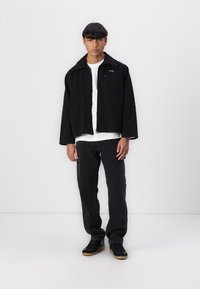 Svart cropped jakke med bred krage, båret over en hvit skjorte. Svart jeans og svarte sneakers fullfører antrekket. Minimalistisk design.