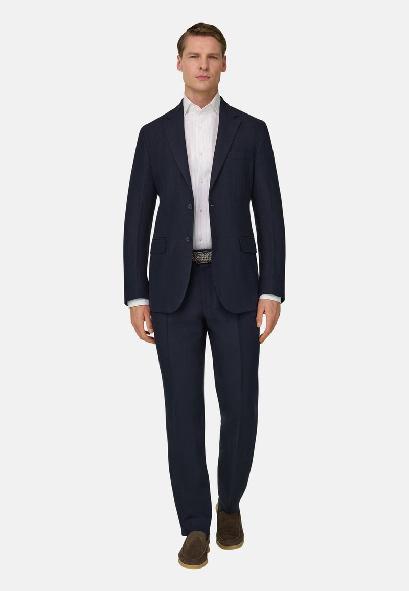 Homme marchant vers l'avant portant un costume bleu marine foncé, une chemise blanche, une ceinture à motifs et des chaussures en daim marron sur un fond clair.