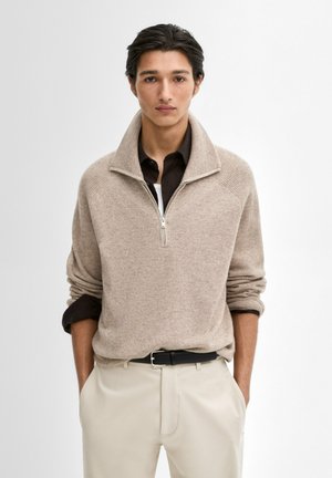 Maglione beige con zip al collo e texture a coste, con spalle scese e zip a metà. Indossato su una camicia marrone scuro e pantaloni chiari.