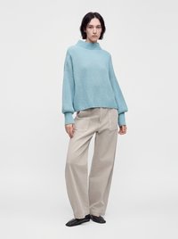 Femme portant un pull côtelé bleu clair, un pantalon beige à jambes larges avec poches avant, et des chaussures plates noires, debout devant un fond clair uni.